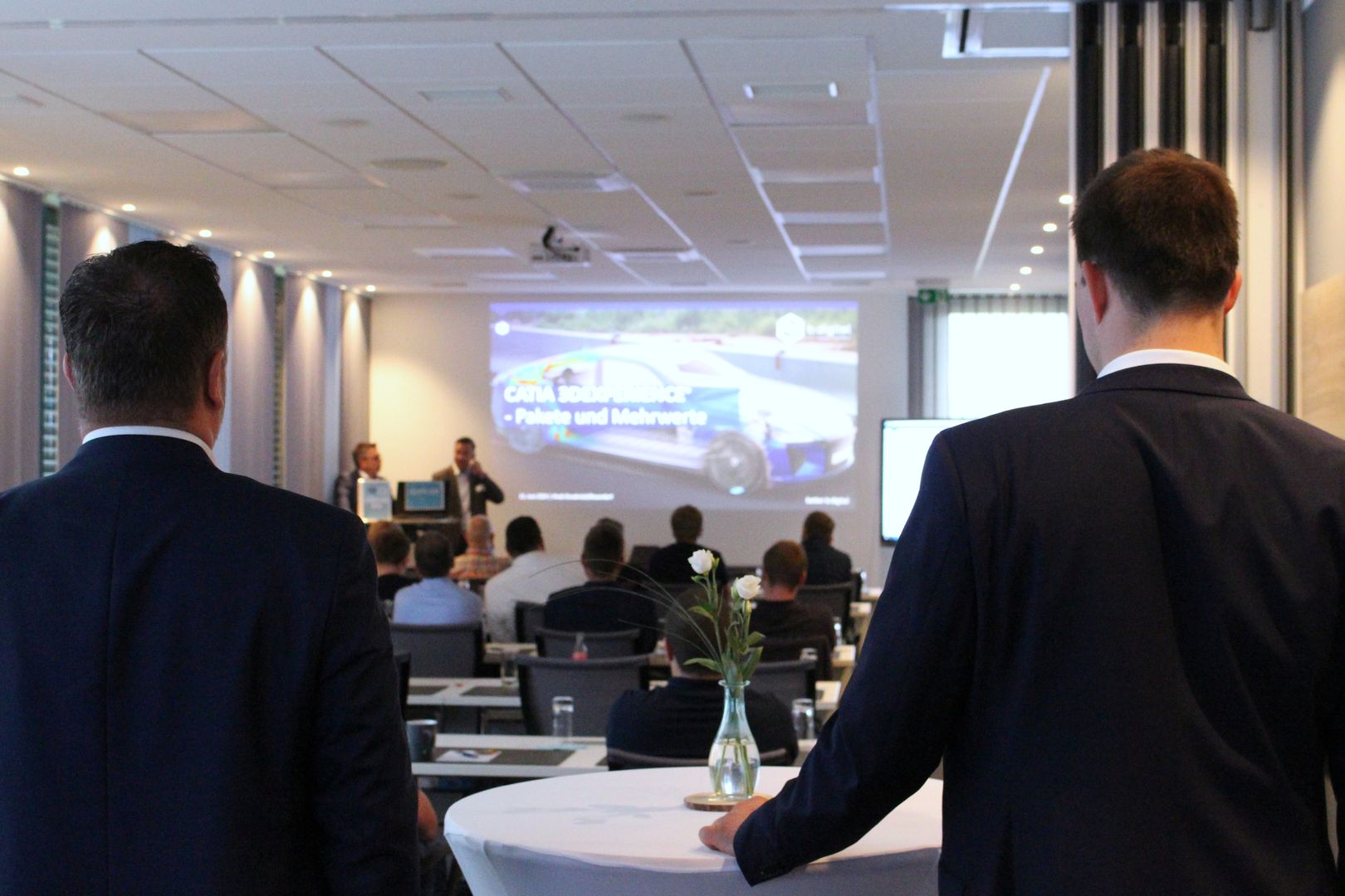 Geschäftsleute besuchen ein Seminar und hören Rednern in Anzügen zu, die auf einem Bildschirm eine Präsentation über Fahrzeuge und Technologie zeigen.